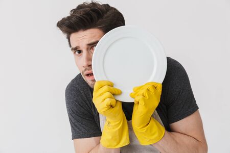 Photo of optimistic young man wearing yellow rubber gloves for hands protection washing dishes while cleaning house isolated over white backgroundの写真素材