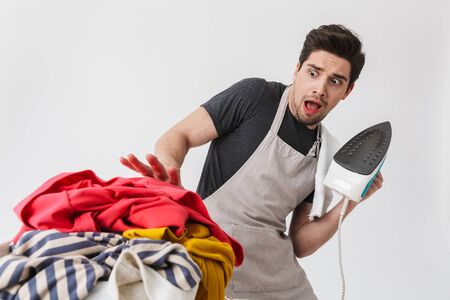 Image of a young scared confused houseman househusband holding iron looking at clothes isolated over white wall background.の写真素材