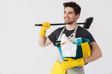 Handsome brunette houseman wearing apron standing isolated over white background, holding bucket full of detergentsの写真素材