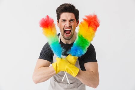 Handsome brunette houseman wearing apron standing isolated over white background, using dust cleanerの写真素材