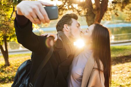 Image of a happy young beautiful loving couple posing walking outdoors in park nature take selfie by mobile phone kissing.の写真素材