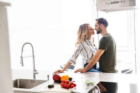 Photo of a tender young romantic loving couple indoors at the kitchen cooking vegetable salad have a breakfast hugging kissing.の写真素材