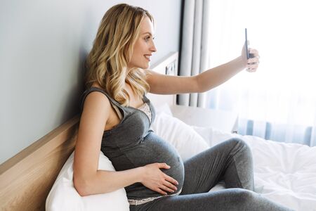 Attractive young pregnant blonde woman relaxing on bed at home, taking a selfie