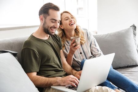 Image of a happy excited young loving couple sitting on sofa at home using laptop computer.の写真素材