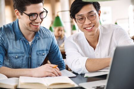 Smart cheerful teenagers studying at the library together, reading books, using laptop computersの写真素材