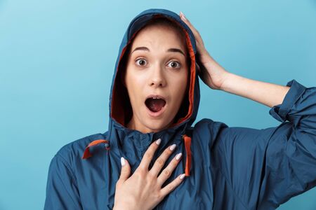 Cheerful young girl wearing raincoat standing isolated over blue background, posing
