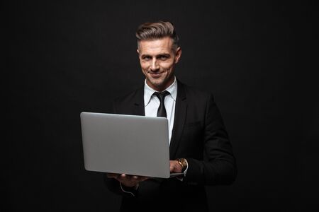 Handsome confident businessman wearing suit standing isolated over black background, working on laptop computerの写真素材