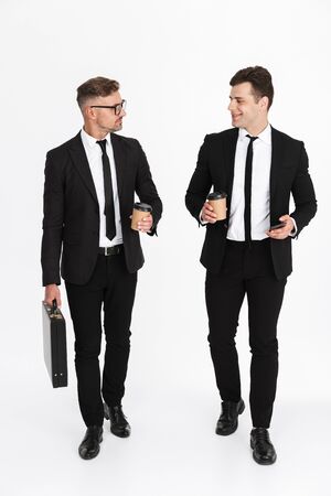 Two attractive smiling confident businessmen wearing suits walking isolated over gray background, drinking takeaway coffeeの写真素材