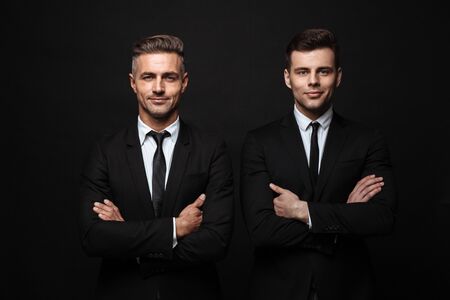 Two confident handsome businessmen wearing suit standing isolated over black background, arms folded