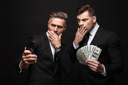 Portrait of shocked two businessmen dressed in formal suit holding cellphone and money banknotes isolated over black wallの写真素材