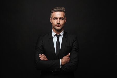 Handsome confident businessman wearing suit standing isolated over black background, arms foldedの写真素材