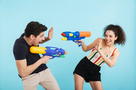 Cheerful young couple standing isolated over blue background, having fun with water guns
