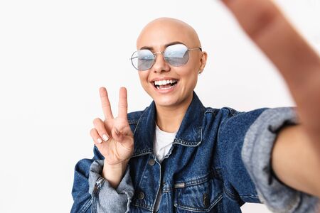 Portrait of a beautiful young hairless woman wearing casual clothes standing isolated over white background, taking a selfie, showing peaceの写真素材