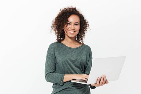 Portrait of a pretty cheerful casual african girl standing isolated over white background, working on laptop computerの写真素材