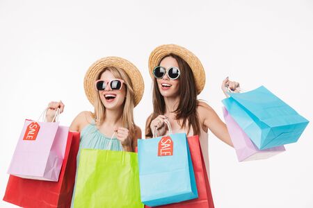 Two cheerful pretty young girls wearing summer hats isolated over white background, shopping together, holding sale bagsの写真素材