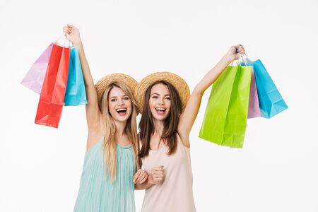 Two cheerful pretty young girls wearing summer hats isolated over white background, shopping together, holding sale bagsの写真素材