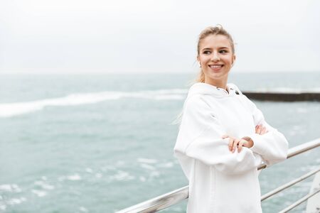 Image of gorgeous joyful woman 20s in white hoodie smiling and listening to music with earpods while...