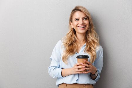 Attractive young blonde businesswoman wearing shirt standing isolated over gray background, holding takeaway coffee cup, looking awayの写真素材