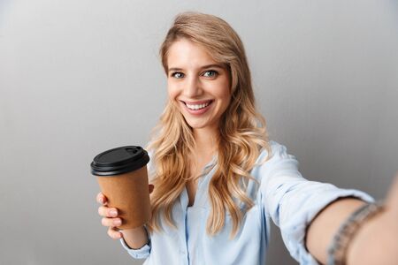 Photo of adorable blond businesswoman smiling and holding paper cup of coffee while taking selfie isolated over gray backgroundの写真素材