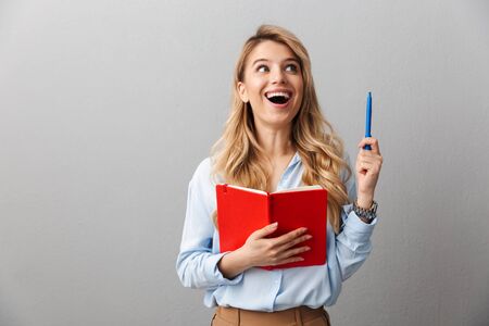 Image of a happy young pretty blonde business woman posing isolated grey wall background writing notes in notebook have an idea.の写真素材