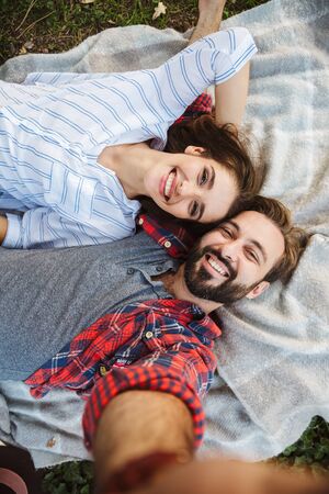 Image of laughing couple man and woman dressed in casual taking selfie photo while lying in green...