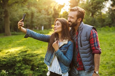 Image of cute couple man and woman dressed in casual wear taking selfie photo on cellphone while walking in green parkの写真素材