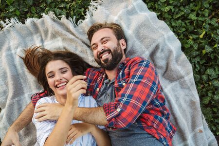 Image of cheerful couple man and woman dressed in casual wear hugging while lying in green parkの写真素材