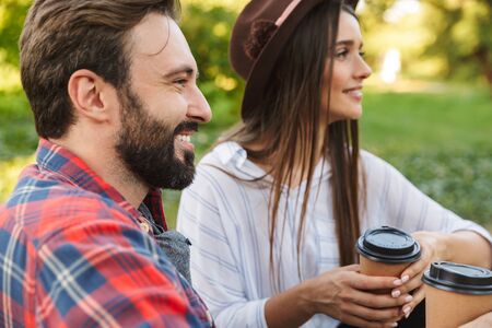 Image of attractive couple man and woman dressed in casual wear drinking takeaway coffee while resting in green parkの写真素材