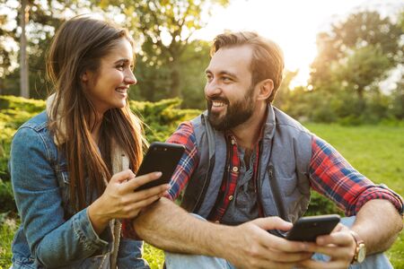 Image of joyful couple man and woman dressed in casual wear using cellphones and looking at each other while resting in green parkの写真素材