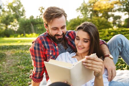 Image of pleased couple man and woman dressed in casual wear reading book while resting in green parkの写真素材