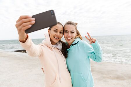 Two happy young fitness women wearing hoodies taking a selfie while stading at the beach, showing...
