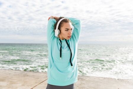 Attractive confident healthy fitness woman wearing headphones exercising outdoors at the beach, stretching handsの写真素材
