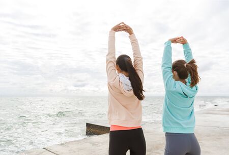 Back view of two attractive young fitness women wearing hoodies exercising outdoors at the seashore, doing stretching exercisesの写真素材