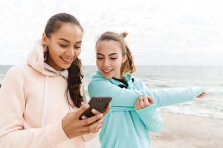 Two pretty smiling fitness women wearing hoodies looking at mobile phone outdoors at the seashore,...