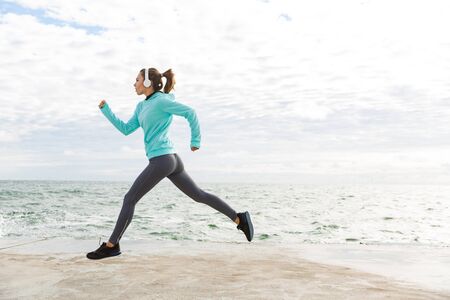 Attractive confident healthy fitness woman wearing headphones exercising outdoors at the beach, runningの写真素材