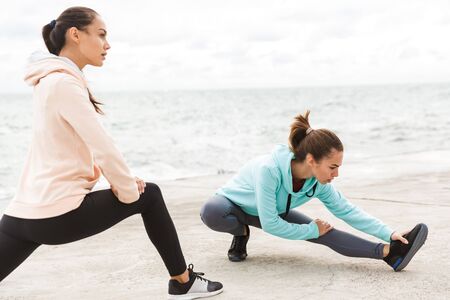 Two attractive confident young fitness women wearing hoodies exercising outdoors at the seashore,...