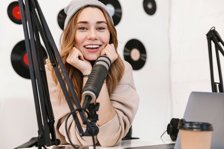 Beautiful happy young female radio host broadcasting in studio, using computer and headphonesの写真素材