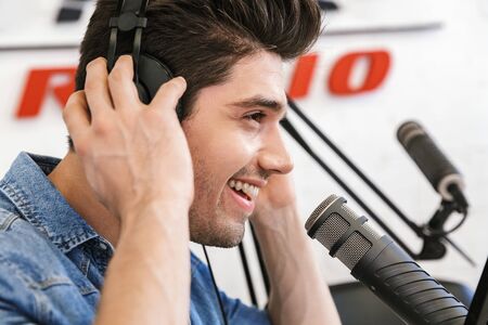 Handsome happy young male radio host broadcasting in studio, using microphone and headphonesの写真素材