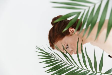 Portrait of a pretty young redhead woman posing isolated over white wall background with leaf green flowers.の写真素材