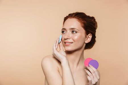 Portrait of a pretty amazing young redhead woman posing isolated over beige wall background holding makeup spongesの写真素材