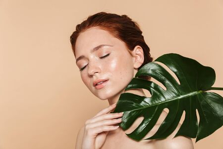 Portrait of a pretty young redhead woman posing isolated over beige wall background with leaf green flowers.の写真素材