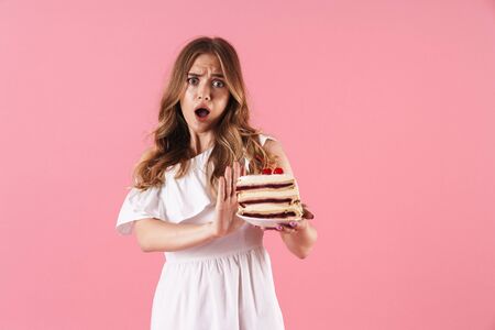 Image of shocked confused woman wearing white dress doing stop gesture and holding piece of cake...