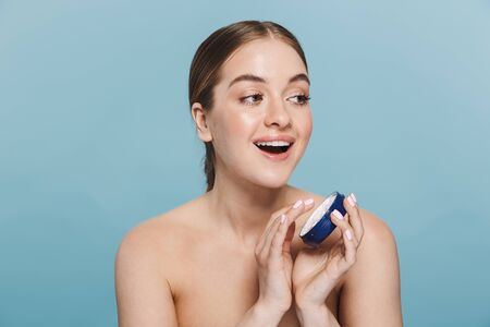 Beauty portrait of an attractive young topless woman isolated over blue background, applying cream...