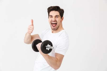 Image of emotional happy excited young man posing isolated over white wall holding dumbbell make exercise make thumbs up gesture.の写真素材