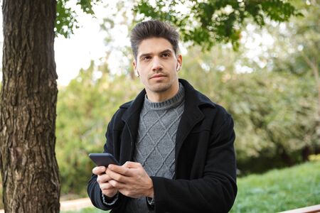 Photo of a serious handsome young man in casual clothing walking outdoors in green park using mobile...