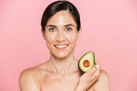 Beauty portrait of an attractive smiling healthy topless brunette woman isolated over pink background, showing sliced avocadoの写真素材