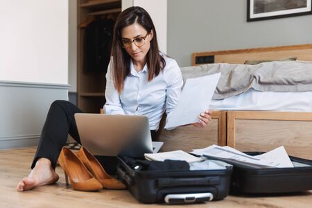 Portrait of beautiful adult businesswoman in formal suit working with paper documents and laptop while sitting on floor near luggage in apartmentの写真素材