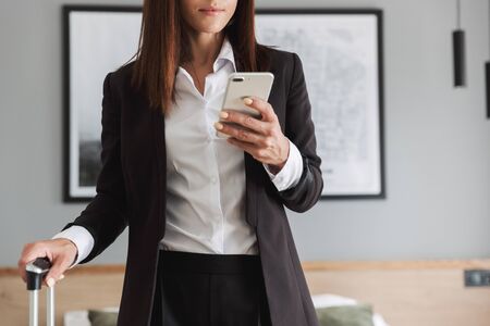 Cropped image of a beautiful young business woman in formal wear clothes indoors at home with suitcase using mobile phone.の写真素材