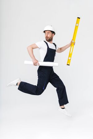 Full length of a confident bearded builder man wearing overalls and hardhat jumping isolated over...