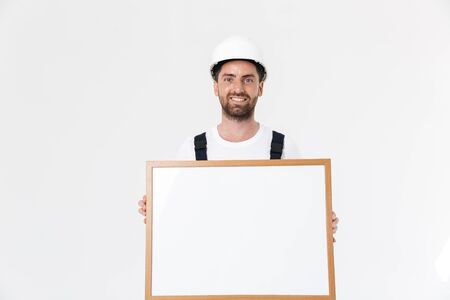 Confident bearded builder man wearing overalls and hardhat standing isolated over white background, showing blank boardの写真素材